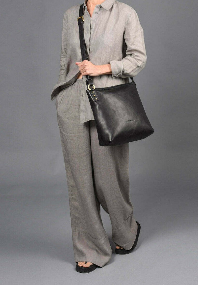 Bolso de hombro de cuero negro con un diseño plano, acentos en hardware dorado y una correa ancha, llevado por una persona vestida con un atuendo de lino gris claro.