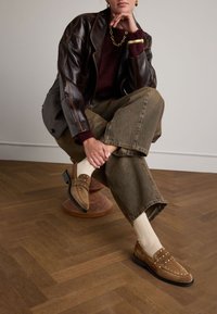 Brown suede loafers with studded accents, paired with cream ribbed socks and wide olive green pants, set against a wooden floor.