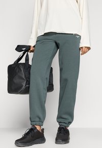 Tumši zaļie sporta bikses ar elastīgām piedurknēm, apvienotas ar melnu sporta somu un melniem sieta sporta apaviem. Apģērbs ir brīvs un vienkāršs.