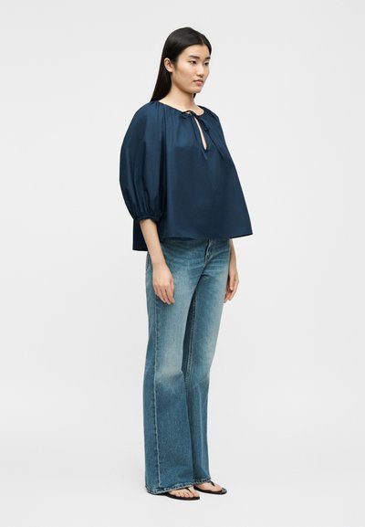 Žena oblečená v voľnej tmavomodrej blúzke s tromištvrte rukávmi, v kombinácii s vyblednutými modrými rozšírenými džínsami a čiernymi sandálmi, stojaca zboku.