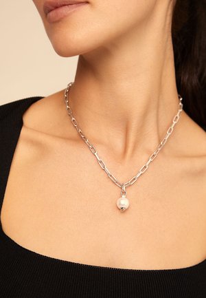 Collier chaîne en argent avec un pendentif unique en grande perle blanche, porté autour du cou d'une femme vêtue d'un haut noir.