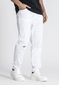 Vaqueros de mezclilla blanca con detalles desgastados, corte recto y bolsillos. Combinados con zapatillas blancas, que tienen un material texturizado y cordones.