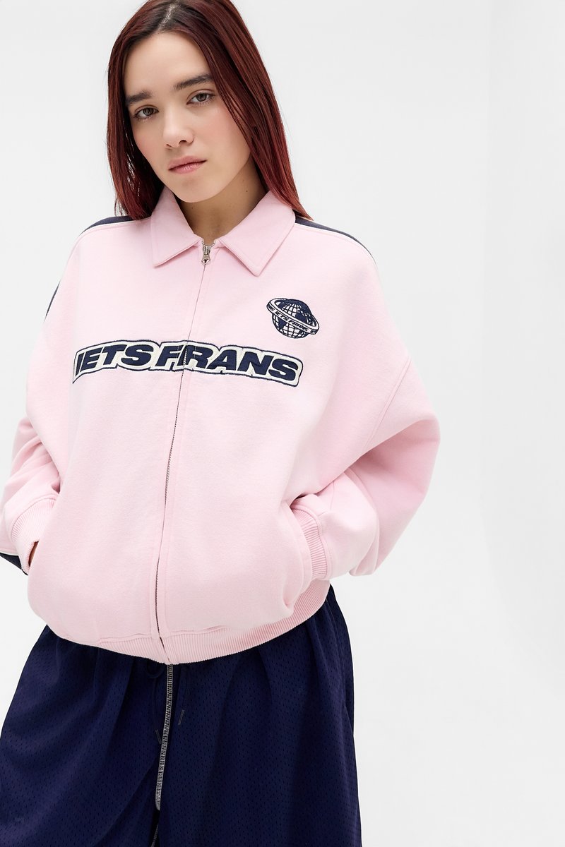 Jeune femme aux cheveux roux lisses portant une veste à zip rose avec l'inscription "Jets Frens" et un logo de globe, associée à un pantalon ample bleu marine.