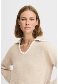 Beige tröja med en scalloped krämfärgad krage och långa ärmar. Tillverkad av mjukt material, med en avslappnad passform som är typisk för vardagskläder.
