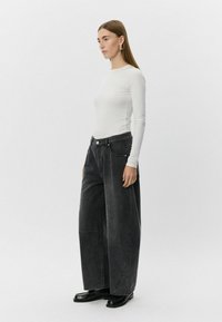 Bela dolga majica z dolgimi rokavi v kombinaciji z oversized črnimi denim hlačami z visokim pasom, sprednjimi žepi in sijajnimi črnimi čevlji.