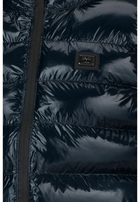 Chaqueta de plumas azul marino con un acabado brillante. Presenta un diseño acolchado horizontal, cierre con cremallera y un parche rectangular con el logo en el pecho.