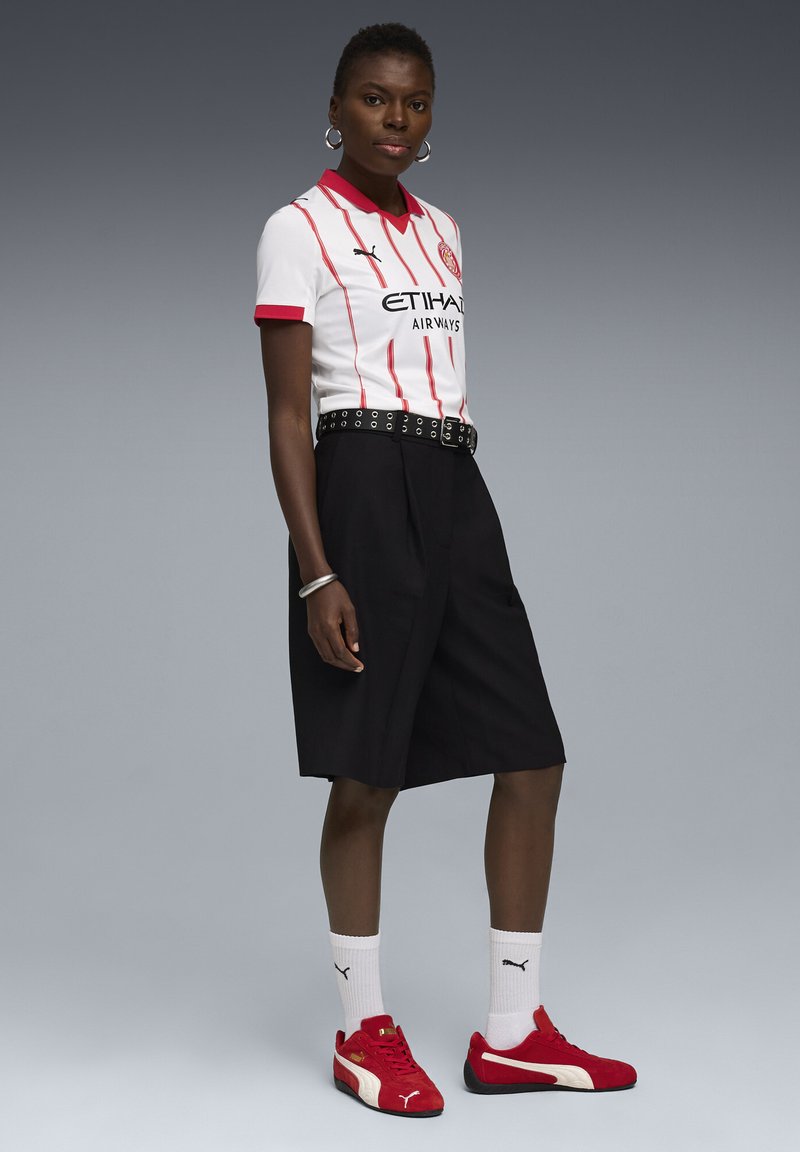 Witte shirt met korte mouwen en rode strepen en kraag, zwarte plooishorts, zwart-witte riem, rode sneakers en witte sokken met logo's.