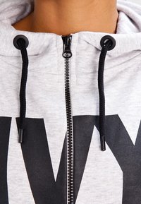 Jasnoszara bluza z kapturem zapinana na zamek błyskawiczny, z czarnymi sznurkami i zamkiem. Na przodzie znajduje się duży czarny nadruk, materiał o miękkiej fakturze i luźnym kroju.