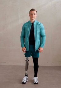 Sportinė apranga, kurią sudaro šviesiai mėlyna džinsinė švarkelis su užtrauktuku, juodas marškinėlis ir žali šortai. Metalinė protezinė koja ir baltos sportbačiai su juodais akcentais.