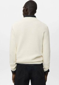 Uomo che indossa un maglione crema a coste lavorato a trama, sopra una camicia nera, visto di spalle su uno sfondo bianco semplice.