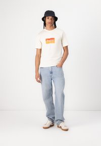 Crème T-shirt met korte mouwen en een oranje grafische print, gecombineerd met lichtblauwe rechte spijkerbroek en witte sneakers met bruine zolen.