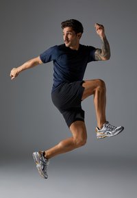Dunkelblaues Sportshirt, schwarze Shorts und silberne Laufschuhe. Die Person ist im Sprung, mit einem sichtbaren Tattoo am Arm.