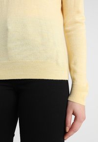 Pull jaune clair à manches longues, en tissu doux, avec un ourlet et des poignets côtelés, porté avec un pantalon noir.