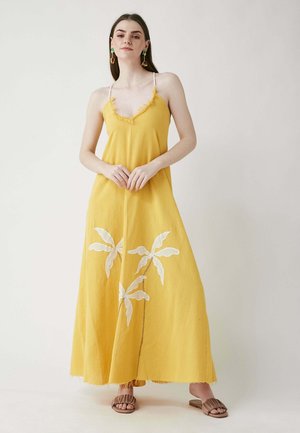 Gelbes Maxikleid mit tiefem V-Ausschnitt, ausgefransten Kanten und weißer Blumenstickerei. Das Model kombinierte es mit braunen Sandalen und bunten Ohrringen.
