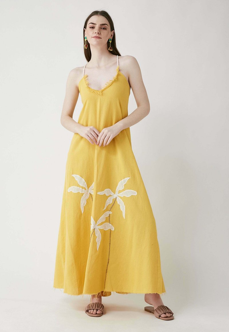 Gelbes Maxikleid mit tiefem V-Ausschnitt, ausgefransten Kanten und weißer Blumenstickerei. Das Model kombinierte es mit braunen Sandalen und bunten Ohrringen.