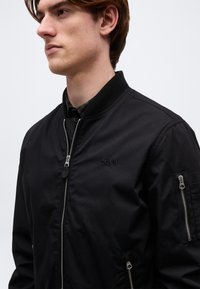 Jeune homme portant une veste bomber noire avec des fermetures éclair à l'avant et sur les manches, faisant face à gauche sur un fond clair uni.