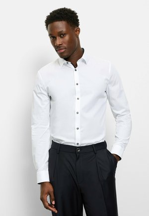 White button-up shirt with a contrasting black collar and buttons, paired with dark trousers. Smooth fabric, long sleeves, and fitted shape.