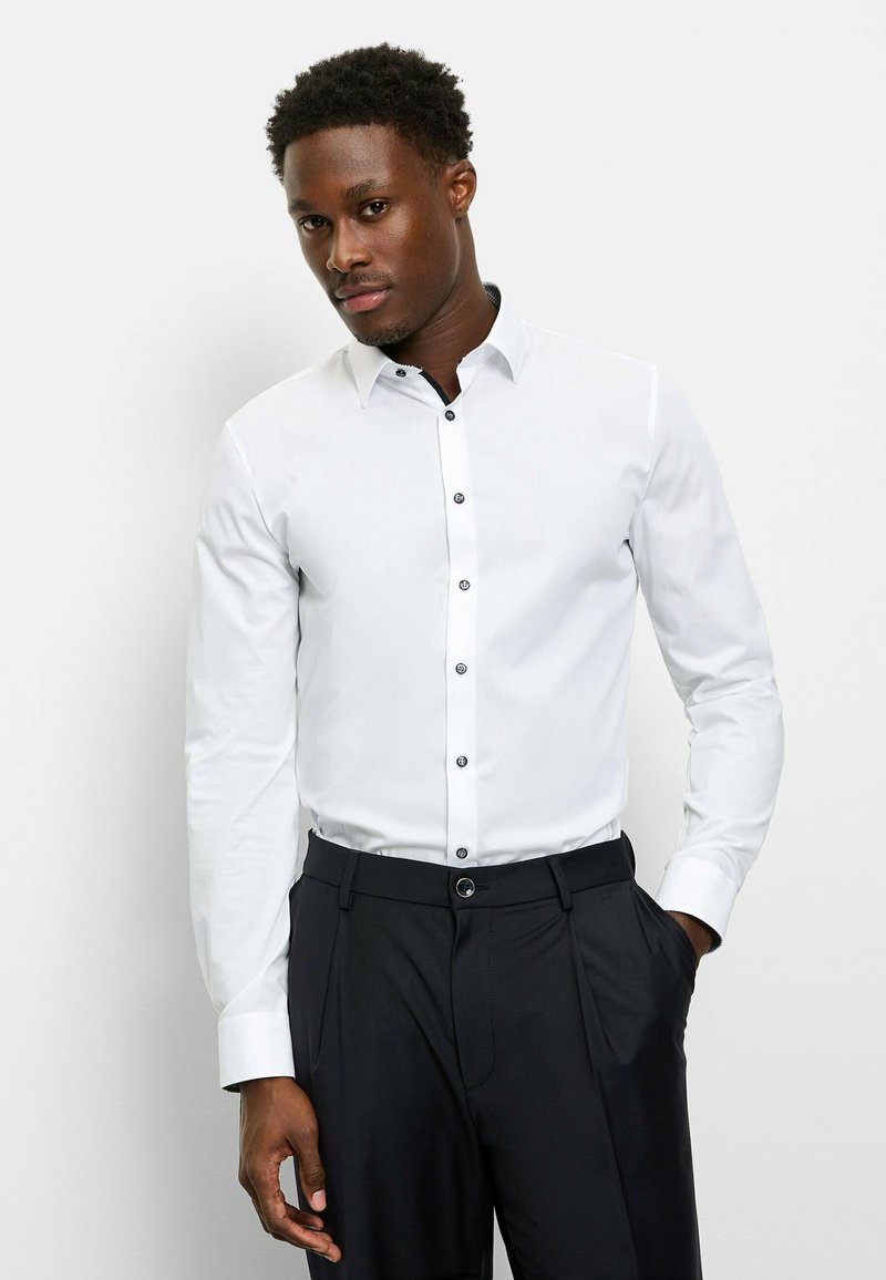 Weißes Hemd mit Knöpfen und einem kontrastierenden schwarzen Kragen sowie Knöpfen, kombiniert mit dunklen Hosen. Glattes Material, lange Ärmel und figurbetonte Passform.