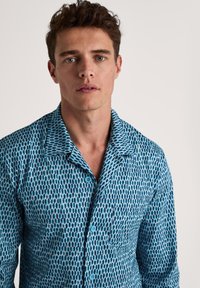 Blauw gestreept shirt met een geometrisch ontwerp, voorzien van een knoopsluiting aan de voorkant, een kraag en een zachte textuur. De stof heeft variërende tinten blauw.