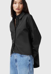 Camicia nera oversize con bottoni, realizzata in tessuto morbido, dotata di collo, maniche ampie e vestibilità comoda, abbinata a jeans azzurri chiari.