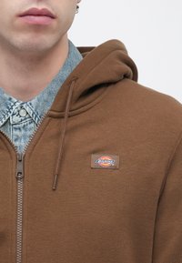 Καφέ φούτερ με φε ρμουάρ και επένδυση στην κουκούλα, με λογότυπο στο στήθος, φορεμένο πάνω από ένα τζιν πουκάμισο με γιακά.