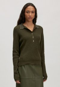 Vrouw in een donkergroene lange mouwen trui met studded kraag en knopen, gecombineerd met een bijpassende donkergroene leren rok voor een effen achtergrond.