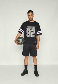 Camisola preta de mangas curtas com "CHAMPION 92" em prata, mangas com listras brancas, combinada com calções pretos e segurando uma bola de basquetebol Spalding.