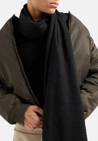 Sciarpa nera realizzata in un tessuto texturizzato, indossata in modo lasco attorno al collo, abbinata a una giacca bomber verde oliva e a una maglietta nera.