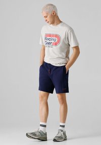 Jonge man met kort blond haar, gekleed in een beige T-shirt met de tekst "Racing Gear", marineblauwe korte broek, witte sokken met zwarte strepen en grijze sneakers, staand.