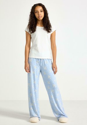 Junge Frau mit langen lockigen Haaren, die ein weißes kurzärmliges Hemd, hellblaue, floral gemusterte, weit geschnittene Hosen und flauschige Hausschuhe trägt und steht.