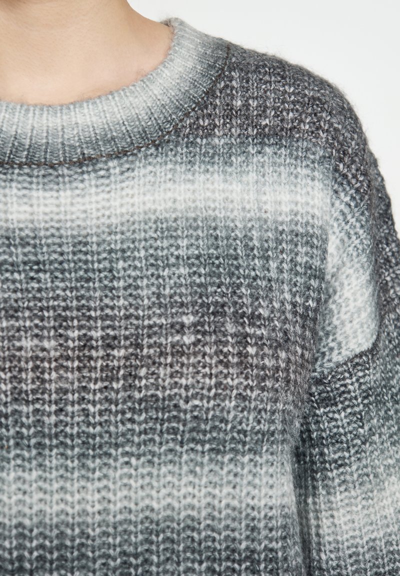 Prim-plan al unei persoane care poartă un pulover din tricot cu nervuri, cu dungi orizontale în nuanțe de gri și alb.