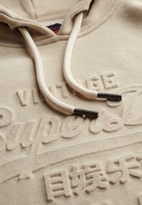 Beige hoodie met verhoogde logo's "Vintage" en "Superdry", voorzien van witte trekkoorden en metalen tips. Zachte textuur en casual ontwerp.