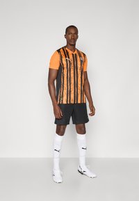 Fodboldtrøje i orange og sort med lodrette striber, parret med sorte shorts og hvide sokker med Puma-logo, båret med fodboldstøvler.