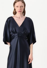 Tamsiai mėlyna satino suknelė su V formos iškirpte, kimono tipo rankovėmis ir susiraukšlėtu juosmeniu. Lygus paviršius, grindims siekiantis dizainas, minimalizmo stilius.