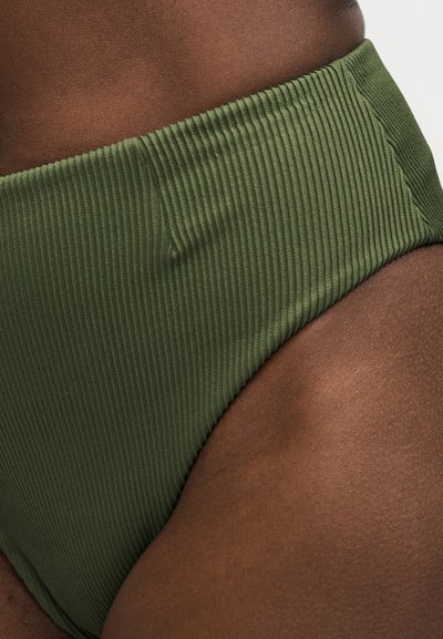 Gros plan sur une peau foncée portant un bas de maillot de bain taille haute en tissu côtelé vert olive sur fond clair.