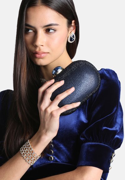 Jeune femme en robe en velours bleu tenant une pochette noire texturée près de son visage, portant un bracelet à perles et des boucles d’oreilles assorties.