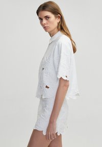 Blusa bianca ricamata con maniche corte e ritagli floreali decorativi, abbinata a una gonna bianca coordinata con bordi scallop.