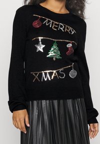 Schwarzer Pullover mit "MERRY XMAS" aus metallischem Faden. Dekoriert mit glitzernden Ornamenten: roter Ball, silberner Stern, grüner Baum und roter Strumpf.