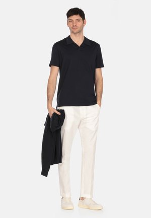 Polo shirt navy, maniche corte, tessuto morbido. Pantaloni bianchi su misura, vestibilità slim. Modello tiene un maglione scuro e indossa scarpe chiare. Sfondo neutro.