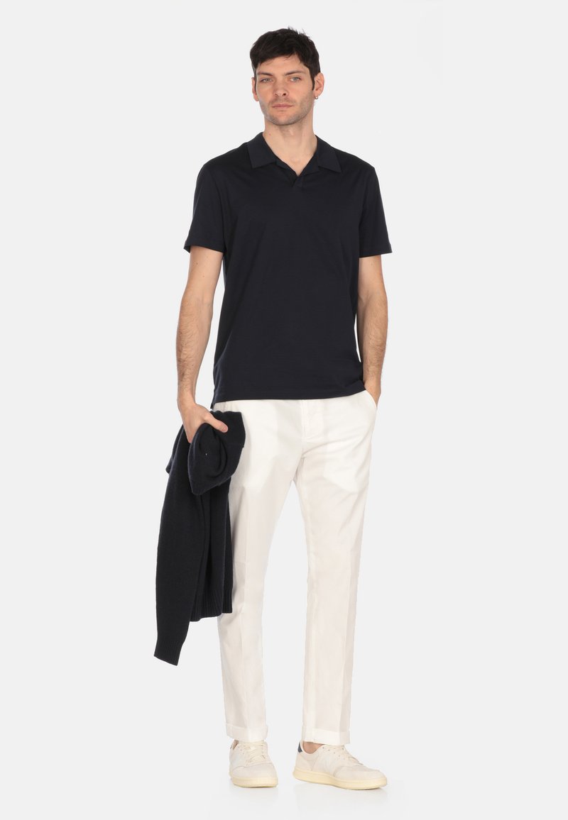 Polo shirt navy, maniche corte, tessuto morbido. Pantaloni bianchi su misura, vestibilità slim. Modello tiene un maglione scuro e indossa scarpe chiare. Sfondo neutro.