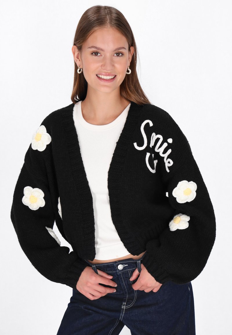 Juodas trumpas kardiganas su baltu užrašu "Šypsena" ir trimatėmis baltomis gėlėmis ant rankovių. Megztas tekstūras, laisvas siluetas ir raukšlėtos rankovės.