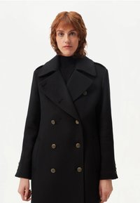 Svart ullkappa med dubbelknäppt front och guldfärgade knappar, bred slagkrage och sidofickor. Mjukt material med skräddarsydd passform.