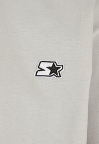 Lys grå bomullsskjorte med en svart-hvit brodert logo med et abstrakt design og stjernedetalj på venstre brystområde.