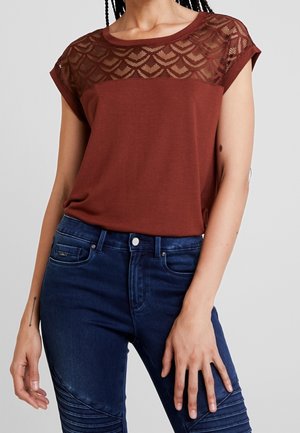 Braunes T-Shirt mit transparentem, gemustertem Spitzeneinsatz und kurzen Ärmeln, kombiniert mit dunkelblauen Jeans mit strukturiertem Design an den Beinen.