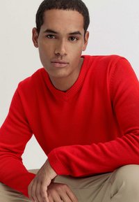 Roter Strickpullover mit V-Ausschnitt, gerippten Bündchen und glatter Textur, kombiniert mit hellen Hosen.