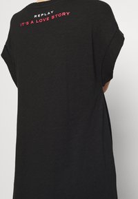 Černé oversized tričko vyrobené z měkké tkaniny. Na zadní straně se nachází bílý a červený potisk s textem "REPLAY IT’S A LOVE STORY."