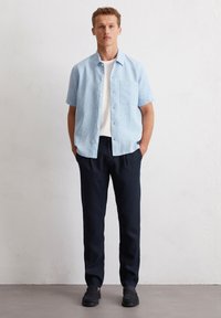Himmelblaues Kurzarmhemd mit einer Brusttasche, kombiniert mit dunkelblauen Hosen und schwarzen Slippern, vor einem strukturierten Hintergrund.