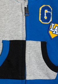 Felpa con zip blu e grigia con accenti neri, dotata di due tasche frontali. Include toppe testurizzate con lettere gialle e blu.