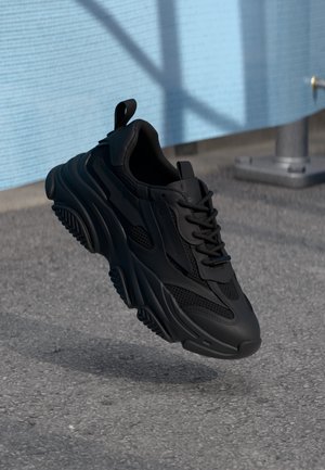 Czarny masywny sneaker z teksturowaną podeszwą i siateczkowymi detalami, pokazany unoszący się nad asfaltową powierzchnią w pobliżu niebieskiej ściany i metalowej instalacji.