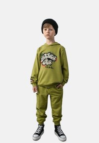 Oliwkowa bluza z kapturem z graficznym nadrukiem, w połączeniu z dopasowanymi dresami i czarnymi wysokimi trampkami. Tekstura wydaje się gładka i casualowa.